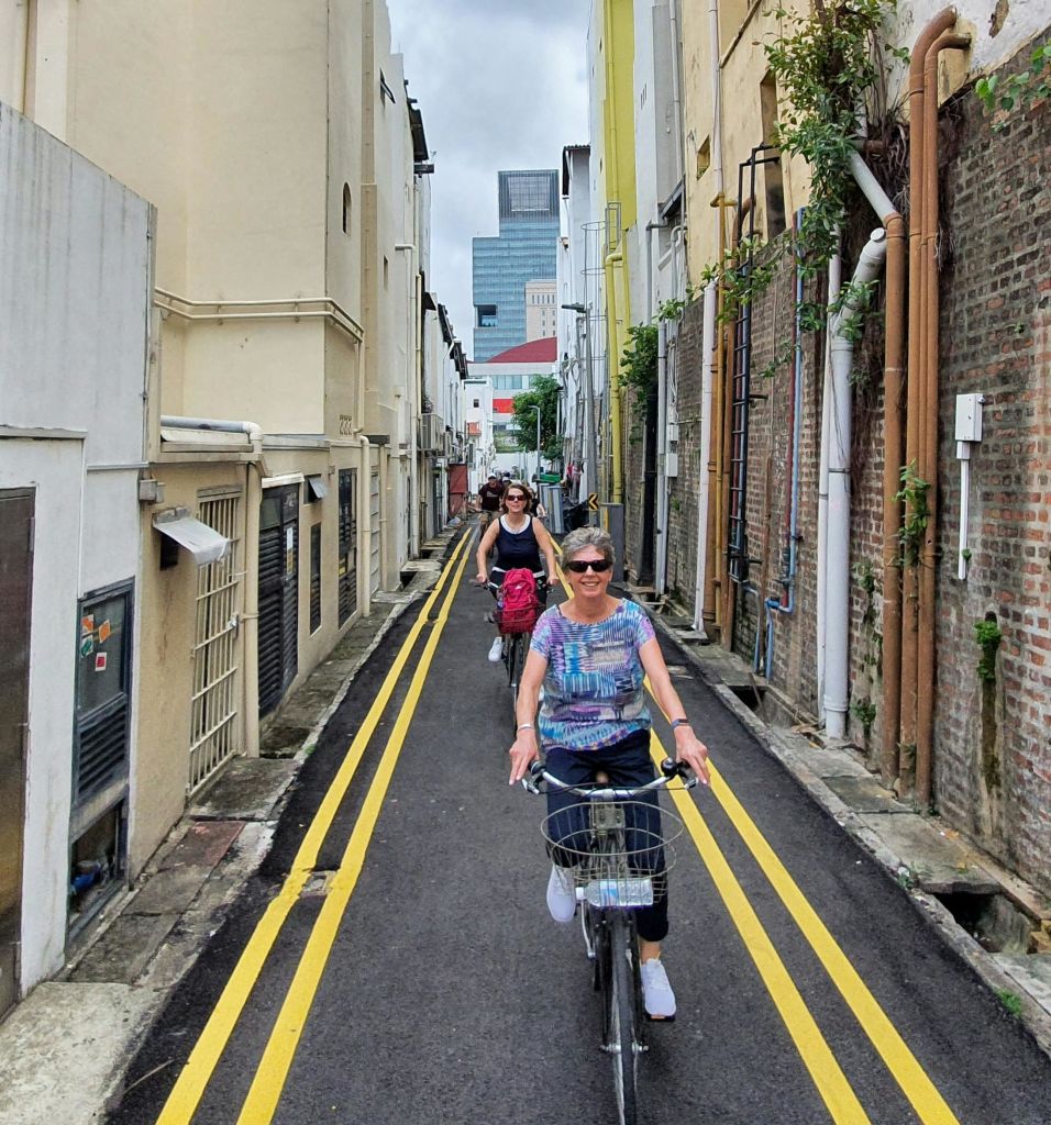 건물, 파이프, 노란색 도로 표시가 줄지어 있는 좁은 골목을 따라 두 사람이 자전거를 타고 내려갑니다. 저 멀리 높은 건물이 보입니다.