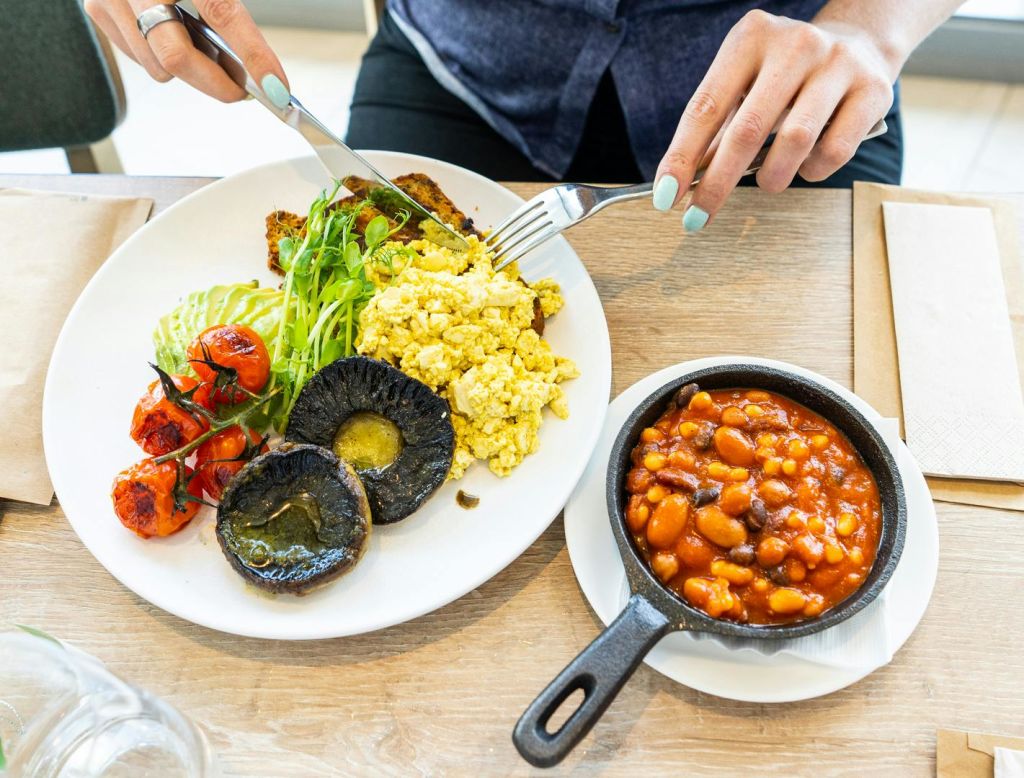 구운 콩의 작은 프라이팬 옆에 스크램블 두부, 구운 토마토, 버섯, 아보카도, 채소가 담긴 접시. 포크와 나이프를 들고 있는 사람.