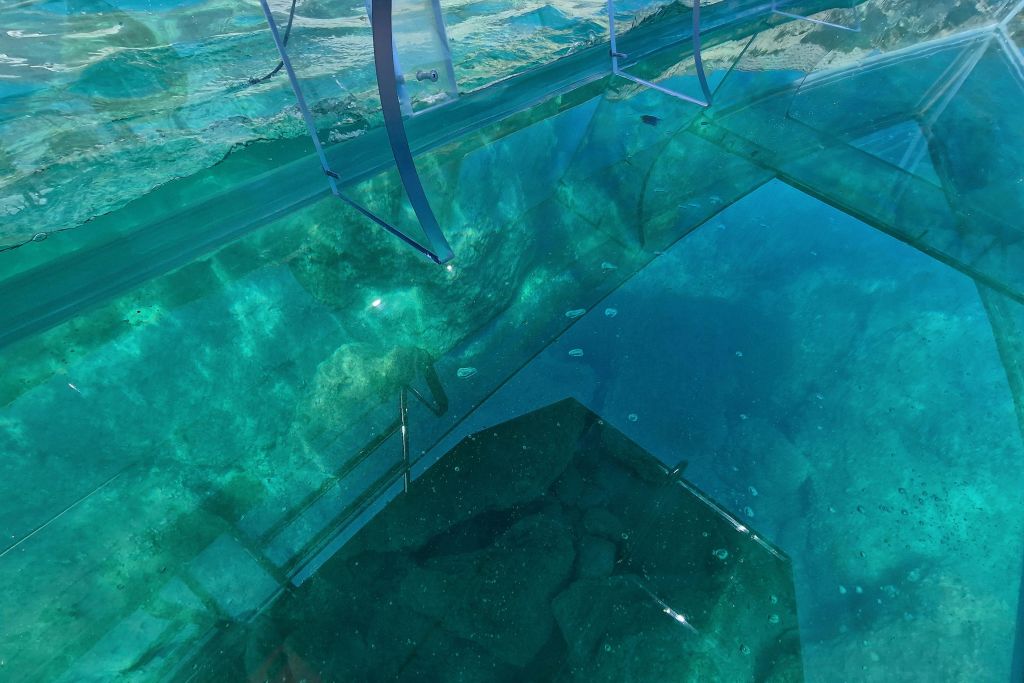 투명한 유리 바닥은 수중 바위와 푸른 녹색 물을 드러내며, 가장자리에는 금속 지지대가 보입니다.