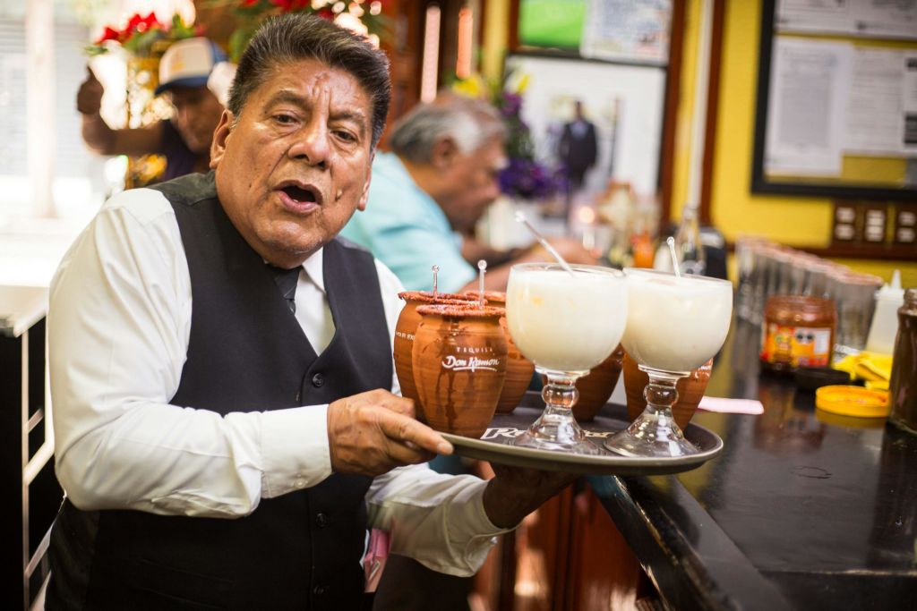 검은색 조끼와 흰색 셔츠를 입은 남성은 음료가 담긴 트레이를 들고 있으며, 다른 남성은 레스토랑을 배경으로 볼 수 있습니다.