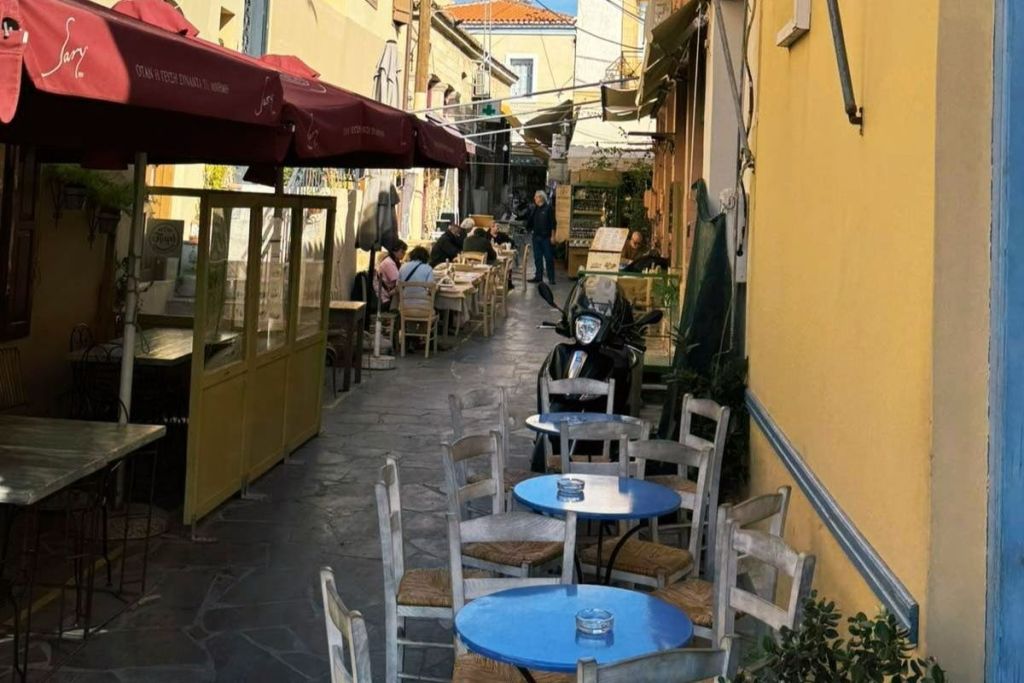 좁은 골목길에 파란색 테이블과 나무 의자가 늘어선 야외 카페. 사람들은 우산 아래 배경에 앉아 있습니다.