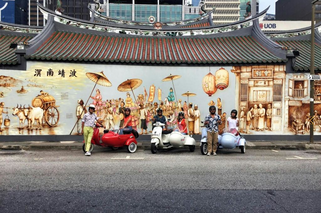 도시를 배경으로 역사적인 장면을 묘사한 화려한 벽화 앞에서 포즈를 취하는 장식용 스쿠터를 탄 사람들.