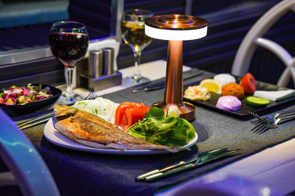 그릴에 구운 생선 요리, 신선한 채소, 음료, 작은 테이블 램프를 갖춘 식당이 있습니다.