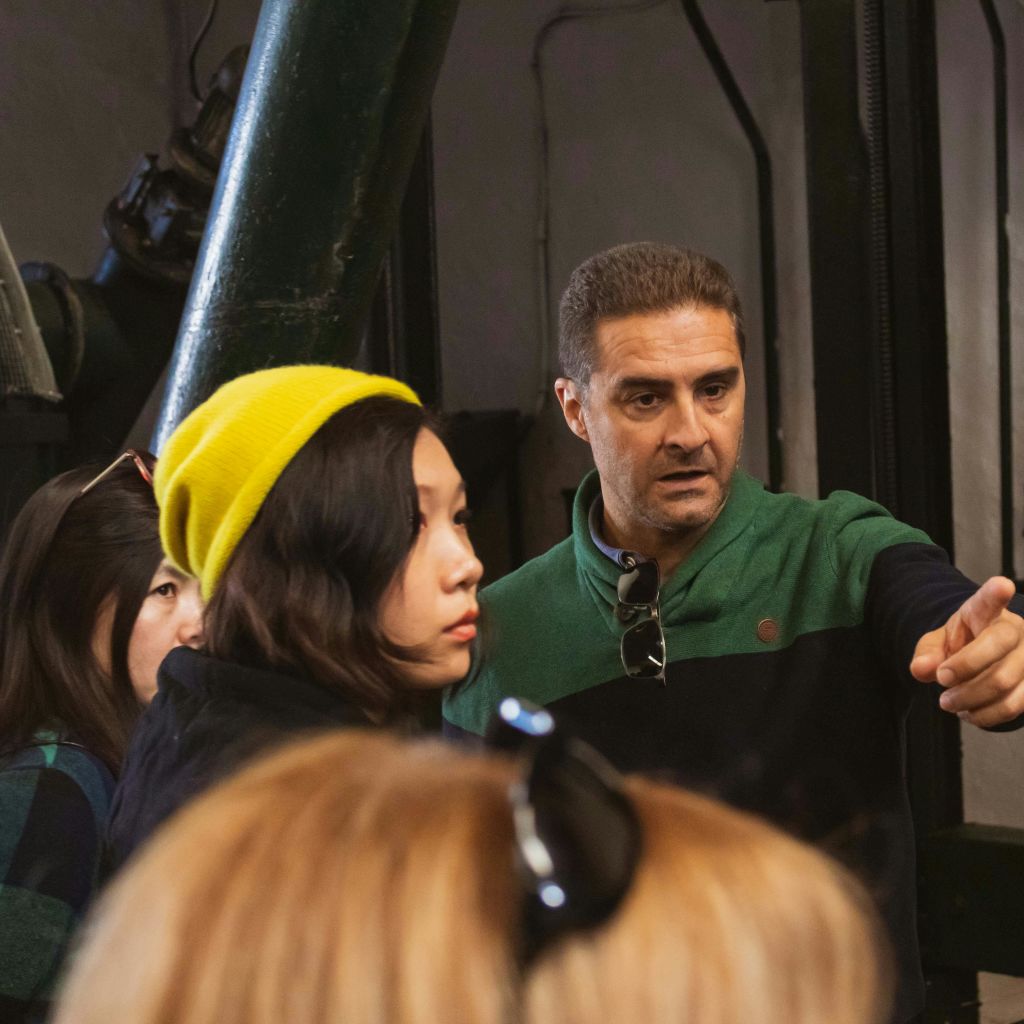 초록색 셔츠를 입은 남성이 실내에서 노란색 모자를 쓴 여성을 포함한 여러 사람들과 대화를 나누며 제스처를 취하고 있습니다.