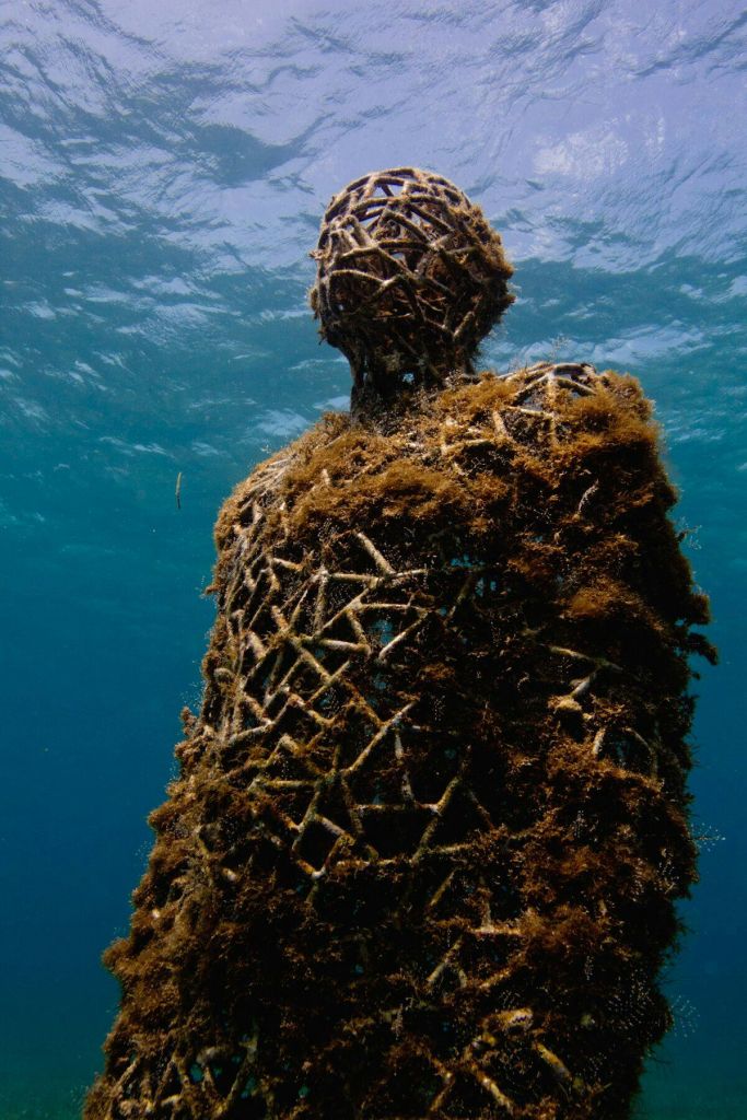 맑고 푸른 바닷물 아래 격자 틀로 만든 해초로 덮인 사람 모양의 잠긴 조각상입니다.