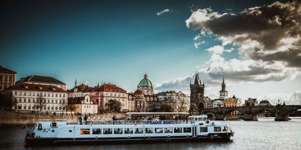 "프라하 보트" 라고 표시된 강 보트가 부분적으로 흐린 하늘 아래 역사적인 건물과 타워를 지나갑니다.