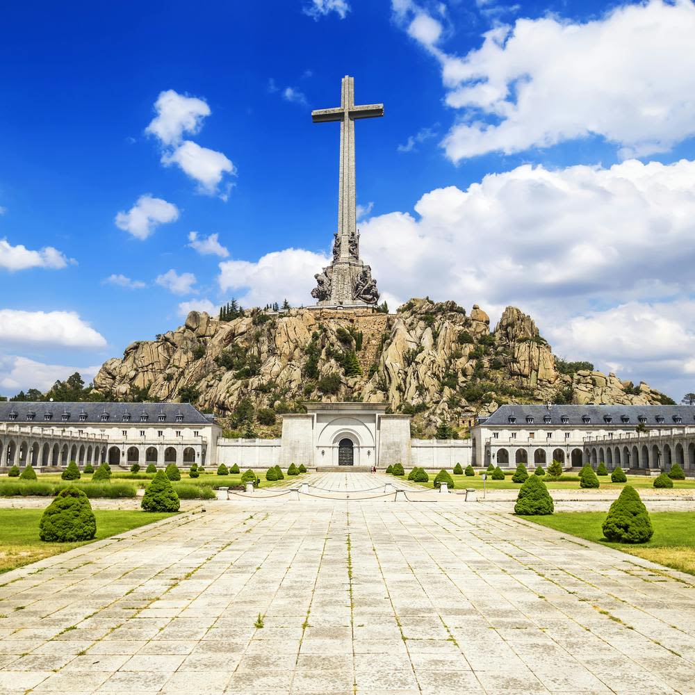 푸른 하늘 아래 수풀을 다듬은 뜰과 기념비적인 건물로 둘러싸인 바위 지형의 언덕 위를 가로지르는 커다란 돌.