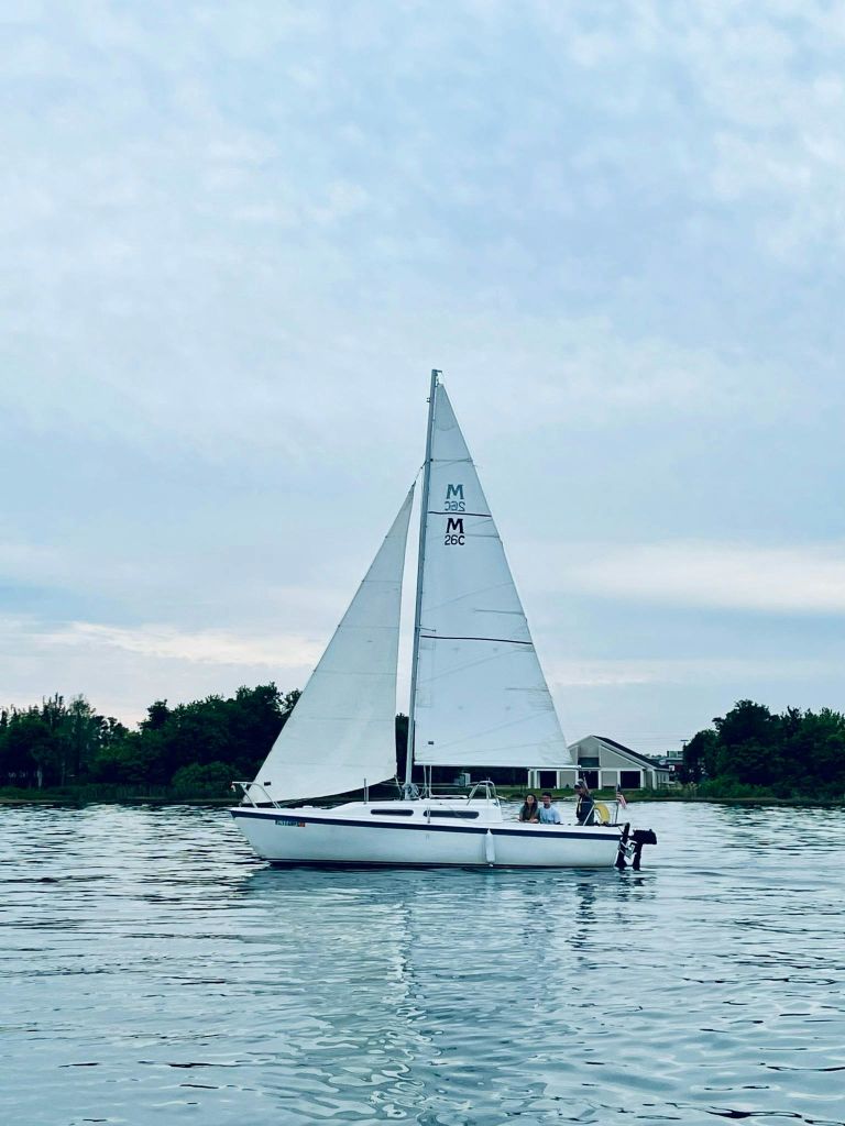 나무와 건물을 배경으로 해변 근처의 잔잔한 물가에서 돛을 올린 작은 흰색 범선.