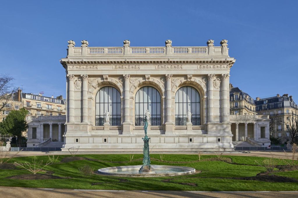 커다란 아치형 창문, 앞에 있는 조각상, 맑고 푸른 하늘 아래 정원에 있는 장식용 분수가 있는 화려한 건물.