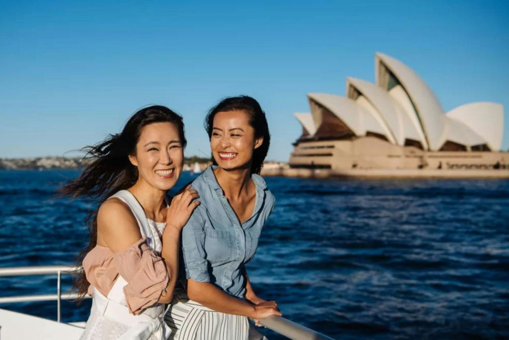 물이 흐르는 보트에서 미소를 짓고 있는 두 여성과 맑고 푸른 하늘 아래 배경으로 펼쳐진 시드니 오페라하우스.