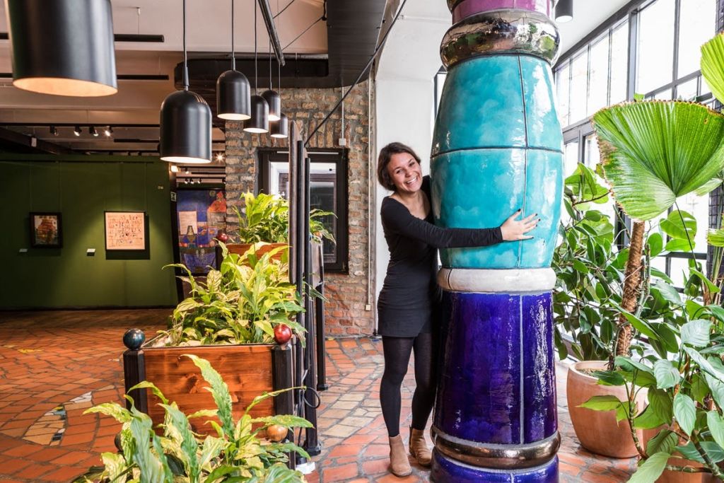 벽돌 벽과 매달린 조명이 있는 식물로 가득 찬 실내 공간에 커다란 다채로운 기둥을 안고 있는 여성.