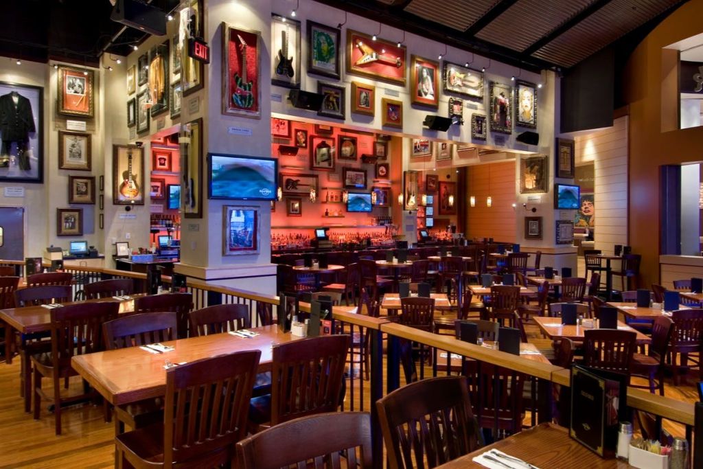 나무 테이블과 의자가 있는 레스토랑 인테리어, 벽에 액자형 기념품, TV, 배경에 바 공간이 있습니다.