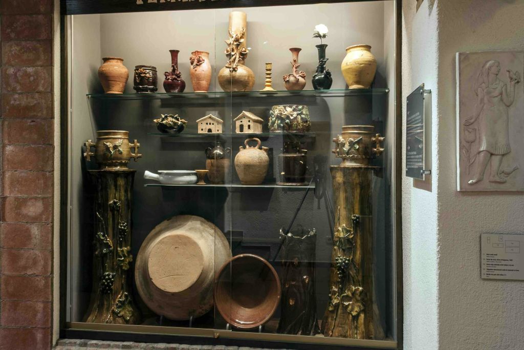 유리 선반에 다양한 세라믹 냄비, 꽃병 및 그릇이 있는 디스플레이 케이스. 바닥에 보이는 커다란 냄비와 장식 기둥.