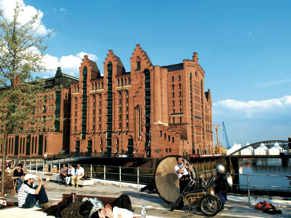 물가에 아치형 창문이 있는 벽돌 건물. 앞쪽에서 한 남성이 난간 근처에서 커다란 북을 연주하고 있다. 사람들이 근처에 앉아 있습니다.