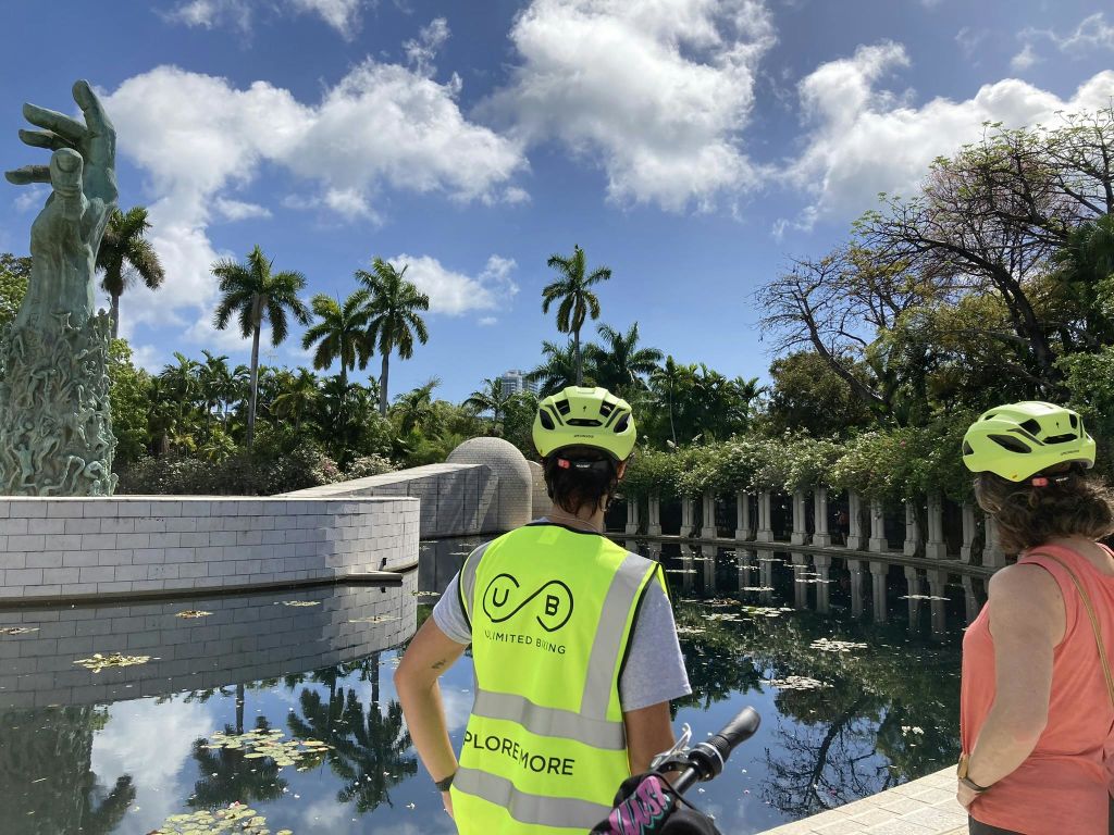 헬멧과 "Unlimited Biking" 조끼를 착용한 사람이 백합 패드, 주변 야자수, 흰색 기둥이 있는 고요한 연못 옆에 서 있습니다.