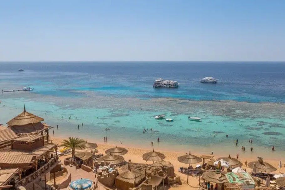 짚 우산이 있는 모래 해변, 물속에 있는 사람들, 푸른 바다에 떠 있는 보트, 머리 위로 펼쳐진 맑은 하늘.