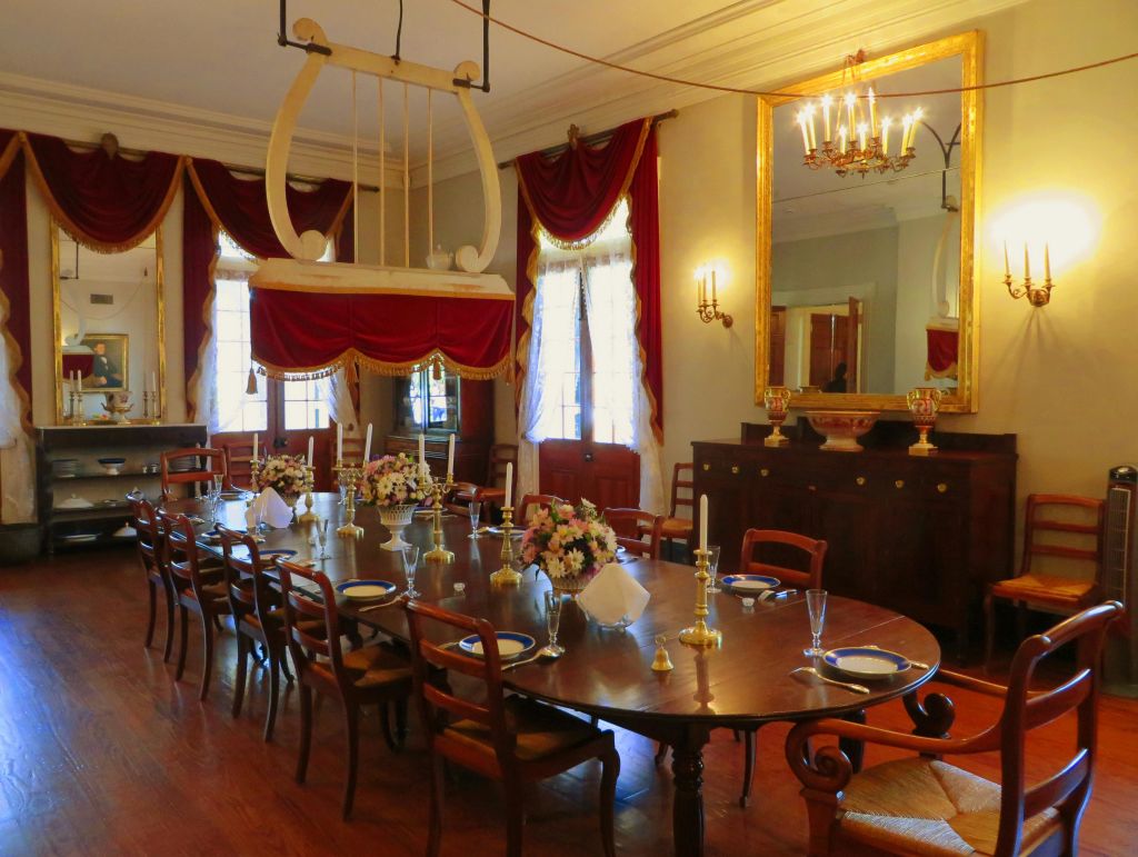 꽃무늬 센터피스, 촛대, 접시가 놓인 긴 나무 테이블이 있는 우아한 다이닝 룸. 빨간색 커튼과 커다란 거울이 인테리어를 돋보이게 합니다.