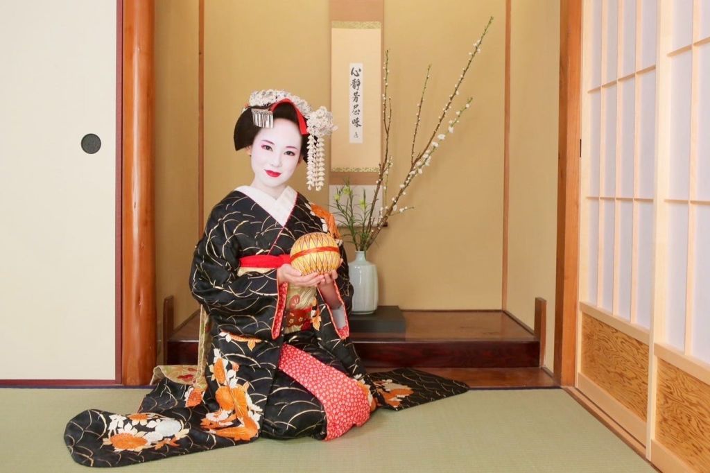 일본 전통 복장을 한 사람이 다다미 위에 앉아 화려한 공을 들고 꽃병과 벽 두루마리를 배경으로 앉아 있습니다.