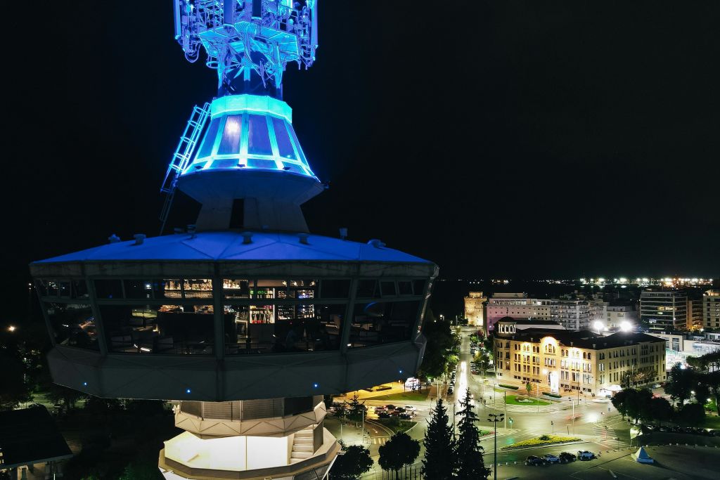 밝게 빛나는 파란색 타워, 주변 조명 건물, 아래로 보이는 거리가 있는 야간 도시 풍경.