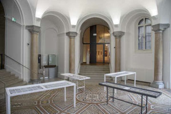 아치형 천장, 기둥 및 문서가 표시된 테이블이 있는 전시 공간의 내부 모습. 계단과 예술작품을 배경으로 진행됩니다.