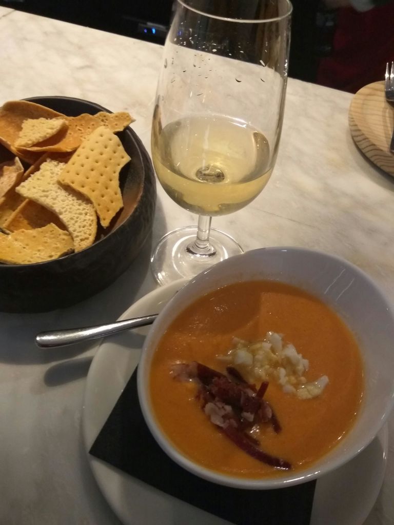 장식이 들어간 오렌지 수프 한 그릇과 화이트 와인 한 잔 옆에 놓인 숟가락, 테이블 위에 놓인 바삭한 빵 조각이 담긴 검은색 그릇.