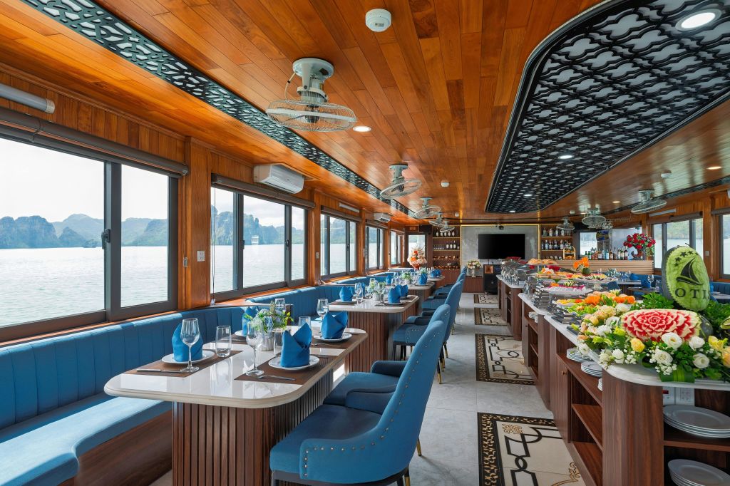 나무 천장이 있는 우아한 보트 식사 공간, 파란색 쿠션이 있는 좌석, 파란색 냅킨이 세팅된 테이블, 뷔페가 마련되어 있습니다. 큰 창문을 통해 바다 전망을 감상할 수 있습니다.