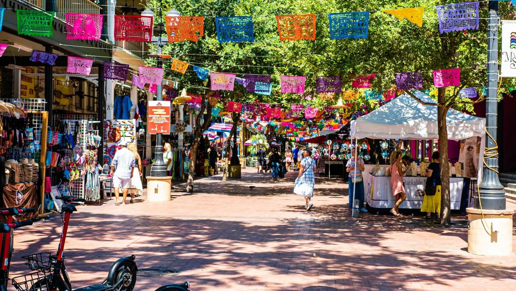 화려한 배너가 걸려 있는 활기찬 야외 시장 풍경, 쇼핑하는 사람들, 나무 아래 다양한 가판대.