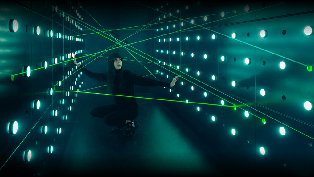 벽에 파란 불빛이 켜진 어두운 방에서 한 사람이 웅크리고 녹색 레이저 광선의 미로를 헤쳐 나갑니다.