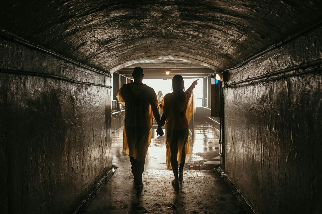 노란 비 판초를 입은 두 사람이 조명이 어두운 터널을 통해 환한 출구 쪽으로 손을 맞잡고 걸어가고 있습니다.