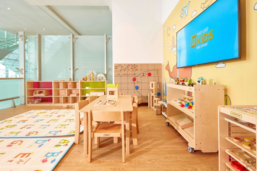 나무 테이블, 의자, 장난감, 선반, "Nat Geo Kids Abu Dhabi" 가 전시된 TV가 있는 다채로운 어린이 놀이방입니다.