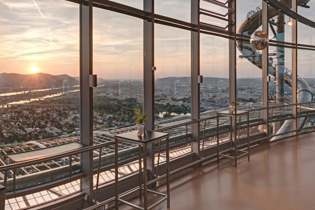 해 질 녘 도시 풍경이 내려다보이는 높은 창문이 있는 현대적인 인테리어로, 창문 근처에 높은 테이블과 스툴이 있습니다.