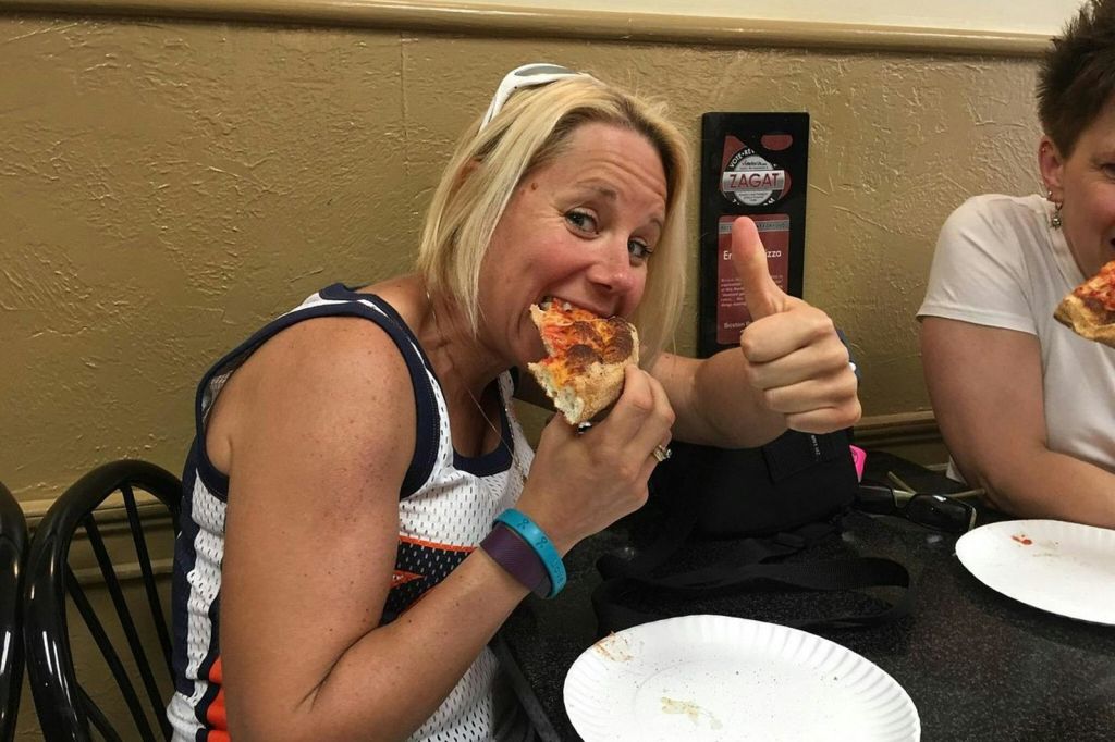 앞에 종이 접시가 놓인 테이블에 앉아 피자를 먹으며 엄지손가락을 치켜세우는 사람.