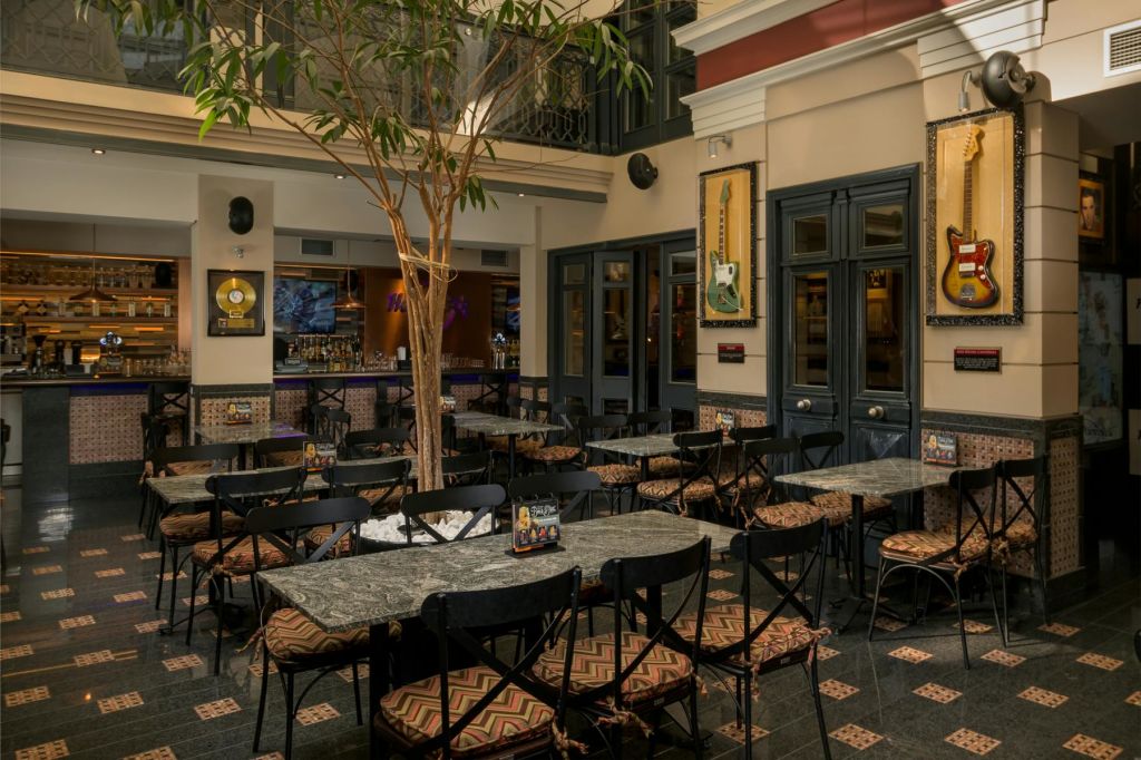 빈 테이블과 의자, 중앙에 나무, 액자에 담긴 음반, 벽에 붙은 기타가 있는 실내 카페. 배경의 바 영역.