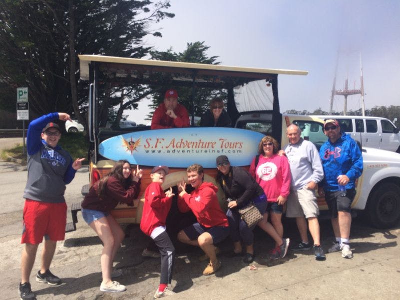 한 무리의 사람들이 주차된 투어 차량 앞에서 "S.F. Adventure Tours" 라고 적힌 표지판을 들고 포즈를 취하고 있습니다.