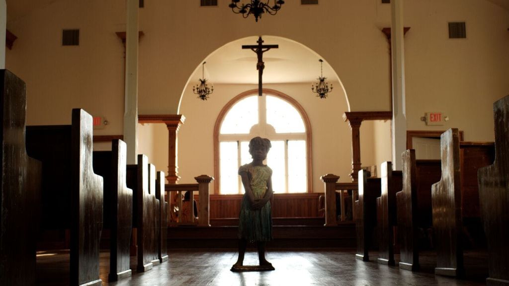 한 어린이가 교회의 중앙 통로에 서 있는데, 양쪽에 퓨즈가 있고, 커다란 십자가가 있으며, 배경에는 아치형 창문이 있습니다.