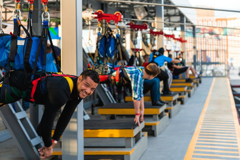 한 남성이 카메라를 보며 미소를 짓고 있는 가운데, 사람들은 짚라인을 탈 준비를 마쳤습니다. 배경은 플랫폼과 장비를 보여줍니다.