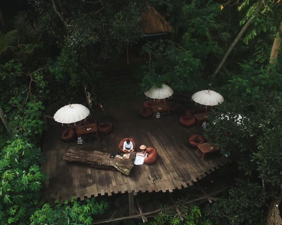 테이블, 의자, 흰색 파라솔, 커다란 나무 테이블 옆에 앉아 있는 두 사람이 있는 숲 속 나무 데크의 공중 전망.