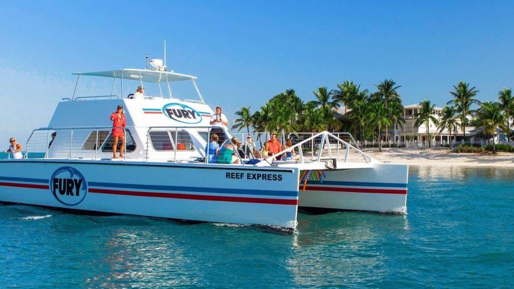 맑고 푸른 하늘 아래 야자수가 있는 모래 해변 근처에 있는 "FURY" 로고가 있는 "REEF EXPRESS" 라는 쌍동선을 탄 사람들.