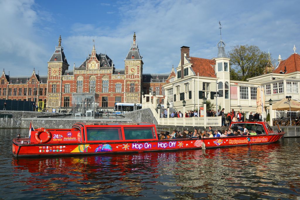 '홉온홉오프 (Hop On Hop Off)' 라고 적힌 붉은 운하 투어 보트가 화려한 건물과 작은 집을 지나고 있으며, 사람들이 탑승하고 있습니다.