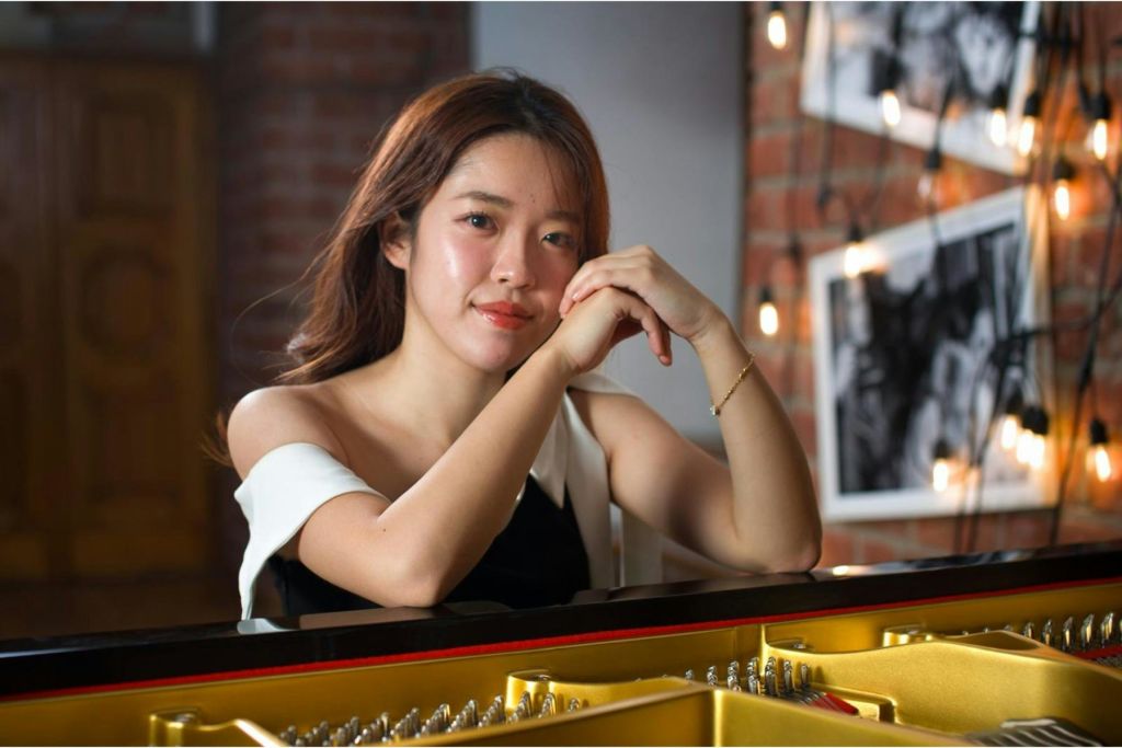 벽돌 벽이 노출된 따뜻하게 빛나는 방에서 그랜드 피아노 옆에 앉아 팔을 얹고 있는 여성.