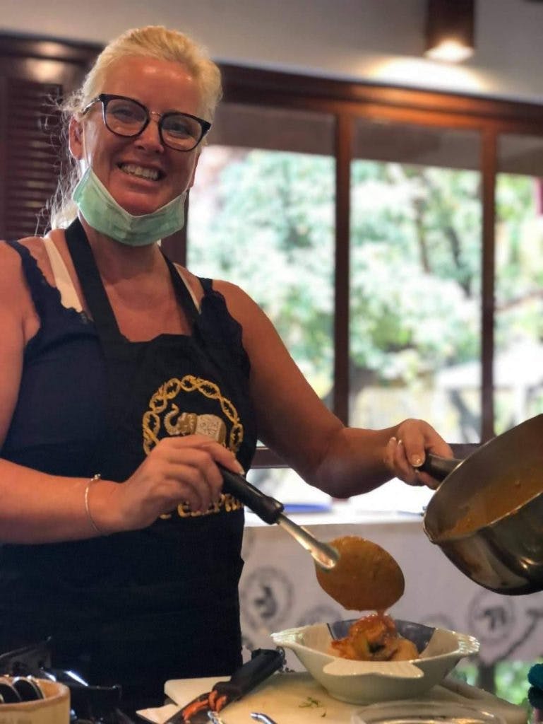 커다란 창문이 있는 주방 환경에서 안경을 쓰고 목에 마스크를 쓰고 팬을 들고 미소를 짓는 사람.