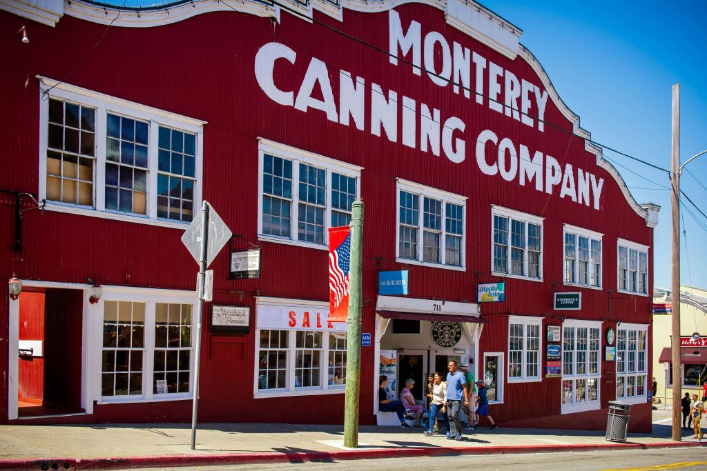 '몬터레이 캐닝 컴퍼니 (Monterey Canning Company)' 라고 적힌 빨간색 건물로 입구를 지나가는 사람들이 있고, 밖에는 미국 국기가 전시되어 있습니다.