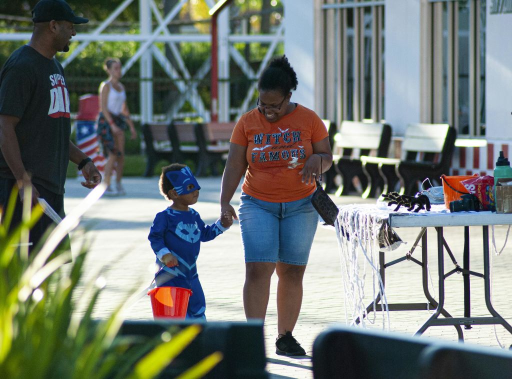 슈퍼히어로 의상을 입은 어린이가 주황색 양동이를 들고 공원에서 주황색 "Witch Famous" 셔츠를 입은 어른과 함께 걷고 있습니다.