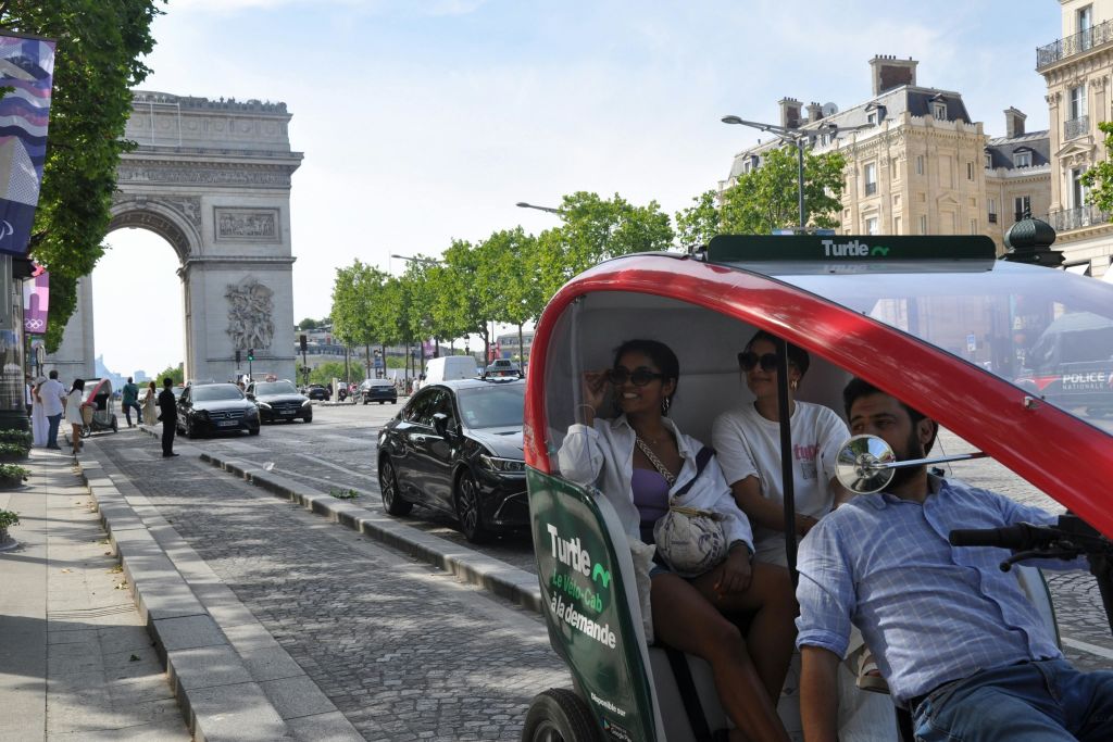도시 거리의 녹색과 흰색 인력거에 세 사람이 앉아 있으며, 배경에는 자동차와 건물이 보입니다.