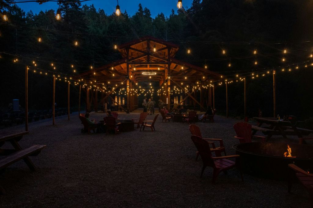 해질 무렵 나무로 둘러싸인 야외 파빌리온에는 줄 조명, 나무 의자, 화덕이 있습니다. 사람들이 안으로 모여듭니다.