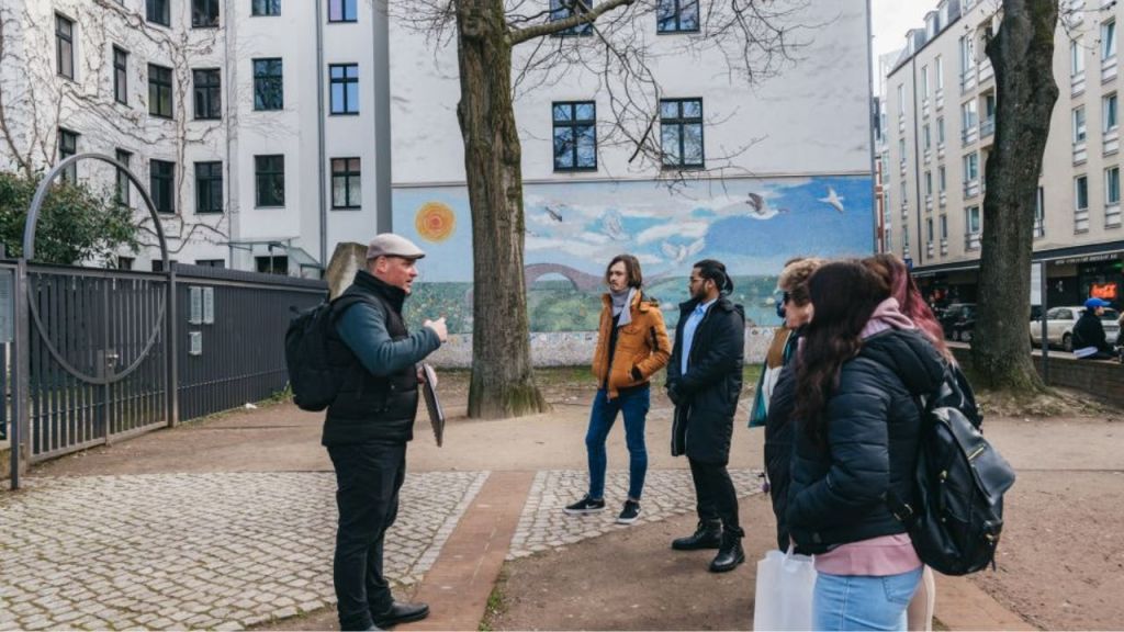 한 무리의 사람들이 나무 근처에 서서 한 사람의 말을 듣고 있습니다. 벽 뒤에 새와 경치가 있는 벽화가 있습니다.