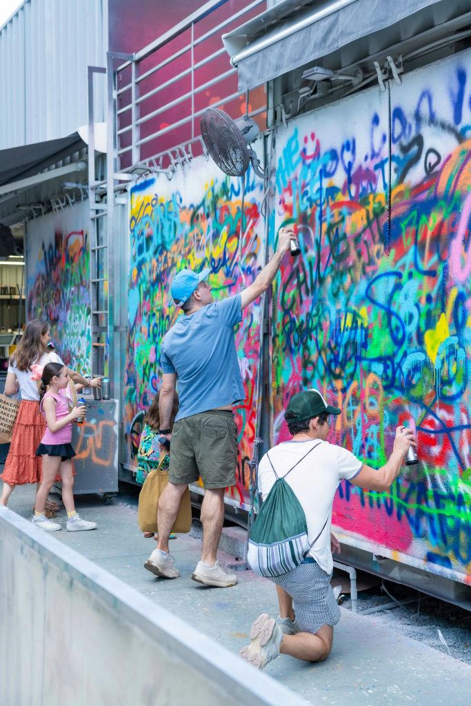 화려한 겹겹이 겹쳐진 디자인으로 생동감 넘치는 그래피티 벽에 스프레이 페인팅을 하는 사람들. 일부 어린이들도 참여하고 있습니다.