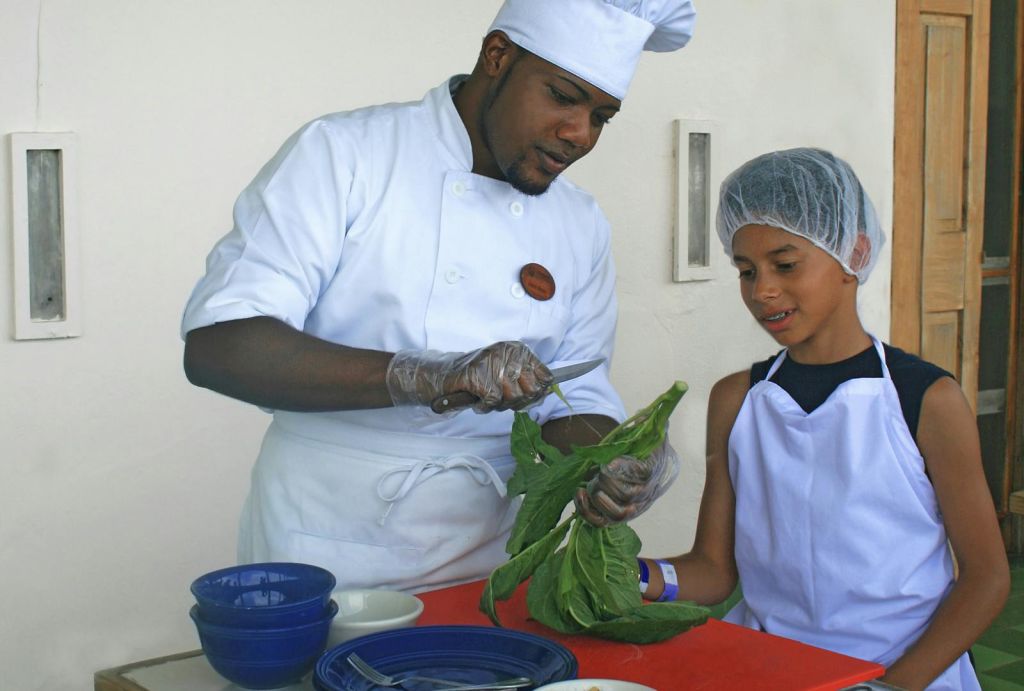 흰색 복장을 한 셰프가 빨간색 카운터 옆에 서서 헤어넷과 앞치마를 두른 어린 소년에게 채소를 준비하는 방법을 보여줍니다.