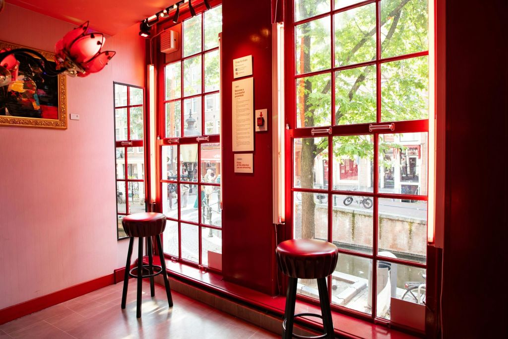 운하와 나무가 내려다보이는 커다란 빨간색 프레임의 창문 옆에는 두 개의 스툴이 놓여 있으며, 밝게 빛나는 내부에는 거울과 벽면 예술품이 있습니다.