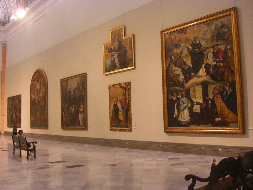 베이지색 벽에 커다란 고전 그림이 전시된 갤러리입니다. 바닥은 대리석이며, 어두운 나무 벤치가 몇 개 있습니다.